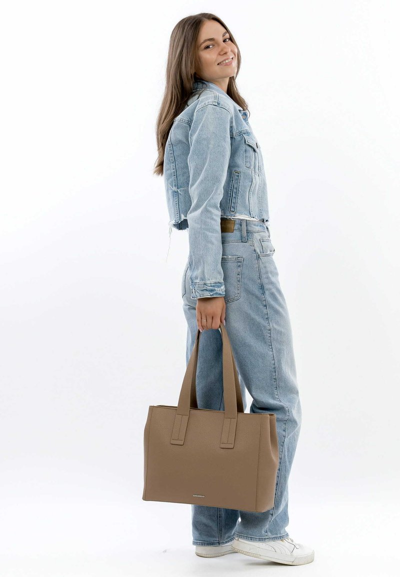 Brun rektangulær totebag med tekstureret overflade, dobbelt robuste håndtag og et diskret logo foran, holdt af en model i denimoutfit.