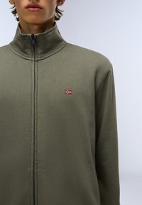 Chaqueta cremallera caqui con alto cuello, hecha de un tejido texturizado. Presenta una pequeña bandera bordada en la parte izquierda del pecho.