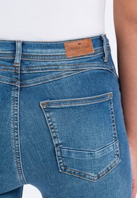 Dżinsy w średnim odcieniu niebieskiego z jasnymi przetarciami, z skórzaną metką na pasku i dwiema tylnymi kieszeniami z pomarańczowymi przeszyciami.