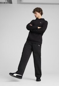 Melna kapuce džemperis un melni bikses no gludas auduma, ar mazu logo uz krūtīm un sānos. Apavi ir melni ar baltām akcentiem.