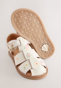 Sandale en cuir blanc avec broderie florale, bout ouvert, trois brides croisées et boucle réglable. Semelle en caoutchouc texturée marron clair avec un détail en forme de coquillage.