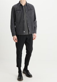 Camicia in denim nero con bottoni automatici e due tasche sul petto, abbinata a pantaloni neri a gamba stretta e scarpe nere. T-shirt grigia sotto.