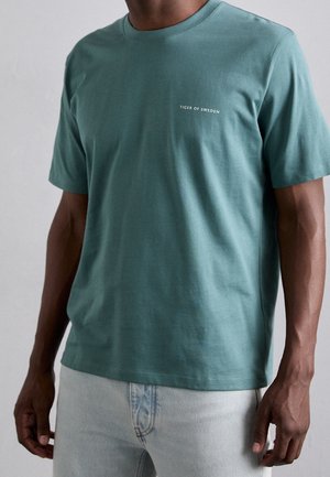 Man draagt een teal kleurige T-shirt met korte mouwen en de tekst "Tiger of Sweden" op de borst, met lichtblauwe spijkerbroek, staand tegen een effen achtergrond.