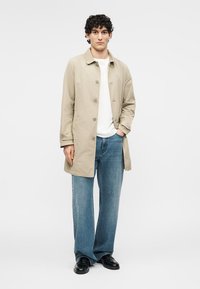 Jaunas vyras dėvi smėlio spalvos užsegamą paltą, baltą marškinėlį, mėlynas džinsus ir juodus batus, stovintis su viena ranka kišenėje ant paprasto balto fono.