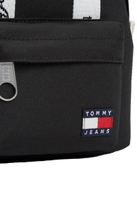 Μαύρη υφασμάτινη τσάντα με υφή στην επιφάνεια, με λευκές και κόκκινες λεπτομέρειες. Περιλαμβάνει ετικέτα με το λογότυπο "TOMMY JEANS" και κλείσιμο με φερμουάρ.