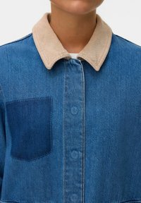 Denim jacket with a light beige corduroy collar, a visible pocket in a darker shade of blue, and a button-up front.
