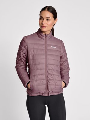 Frau mit dunklen Haaren im Pferdeschwanz trägt eine mauve gesteppte Reißverschlussjacke mit „hummel“-Logo, schwarze Leggings, Hände in den Jackentaschen, schaut seitlich.