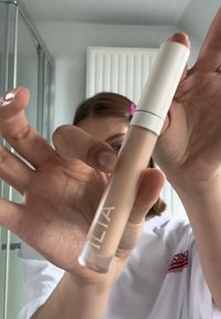 ILIA concealer i en transparent tub med ett vitt lock, har en ljusbeige vätskekonsistens. Varumärket är tryckt i vitt.