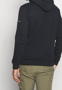 Felpa nera con cappuccio in misto cotone, con tasca a marsupio, polsini a costine e logo in rilievo sulla manica. Abbinata a pantaloni cargo color oliva.