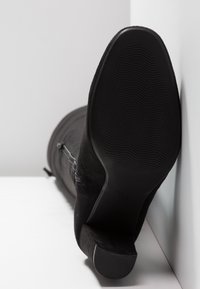 Botín negro de ante hasta el tobillo con tacón ancho, patrón de suela visible y cremallera lateral, posicionado sobre una superficie blanca contra una pared blanca.