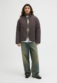 Veste matelassée marron avec col montant et deux poches avant, associée à un jean vert ample et des chaussures foncées, présentée sur un fond neutre.