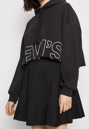 Vrouw draagt een zwarte cropped hoodie met de tekst "LEVI'S" en een zwarte rok met hoge taille, tegen een effen lichte achtergrond.