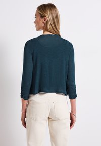Teal beskuren långärmad topp med en mjuk textur och rynkade ärmar, tillsammans med vita högmidjade jeans. En enkel, avslappnad design.