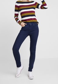Top de manga larga a rayas en burdeos, oliva y azul marino sobre un fondo blanco; combinado con unos jeans oscuros ajustados y zapatillas blancas.