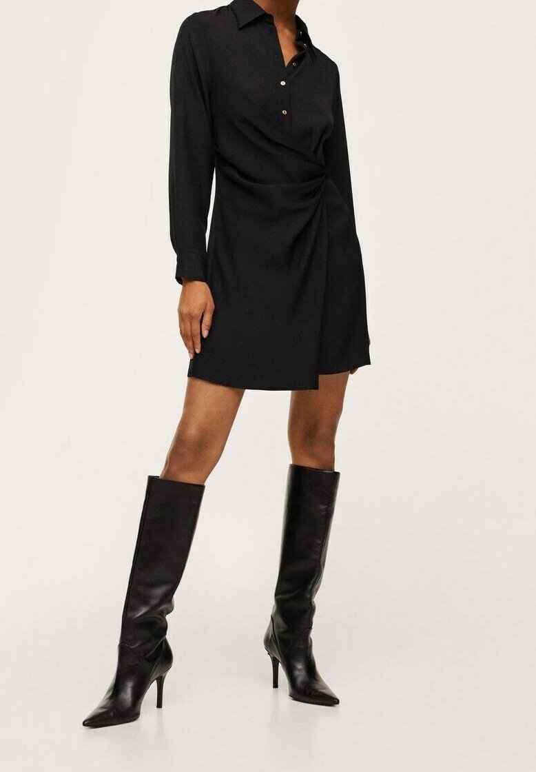 Femme portant une robe chemise noire à manches longues avec un détail drapé sur le devant et des bottes en cuir noir à talons montant jusqu'aux genoux, debout sur un fond blanc.