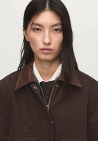 Veste en coton marron avec un col en cuir marron foncé, fermeture éclair argentée et poches latérales, superposée sur une chemise blanche.