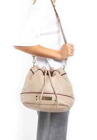 Beige lederen emmer tas met bordeaux accenten, trekkoordsluiting, goudkleurige hardware en een gestructureerd oppervlak.