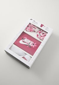 O conjunto Nike de 3 peças inclui um body rosa com detalhes florais, uma faixa para a cabeça combinando e meias brancas com um logotipo. Embalado numa caixa retangular.