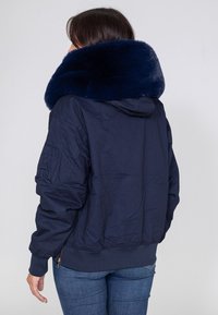 Marineblaue Bomberjacke mit einem plüschigen, gefälschten Pelzkragen. Verfügt über elastische Bündchen, eine Reißverschlusstasche am Ärmel und einen taillierten Bund.