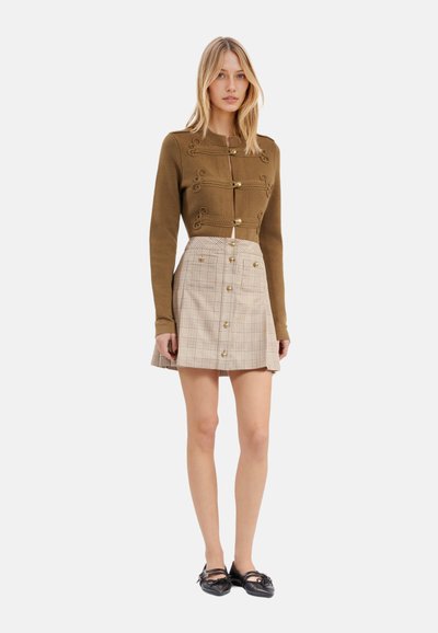 Olijfgroene cropped sweater met decoratieve gouden knopen en borduursels, gecombineerd met een beige geruite minirok met frontknopen. Zwarte schoenen.