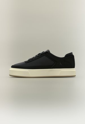 Sneaker nero basso con suola spessa bianca, allacciatura frontale e sottile logo "cK" sul lato.