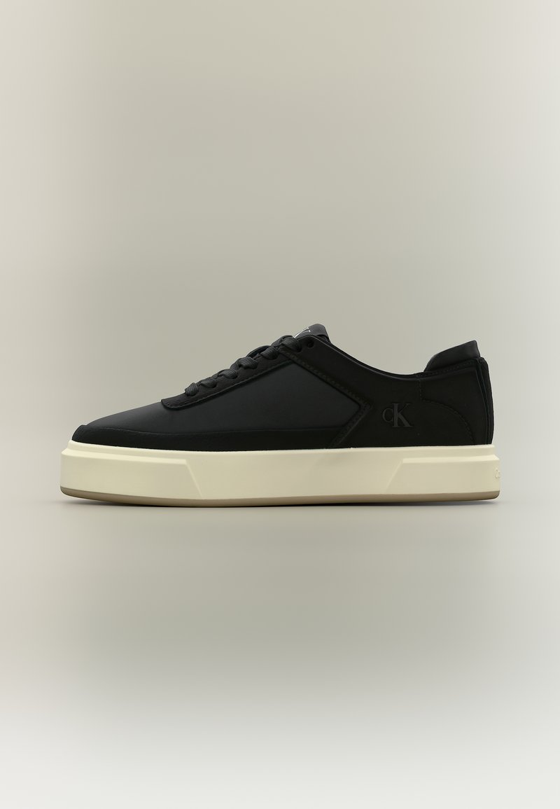Μαύρο αθλητικό παπούτσι χαμηλού ύψους με λευκή χοντρή σόλα, κορδόνια στο μπροστινό μέρος και διακριτικό λογότυπο "cK" στο πλάι.