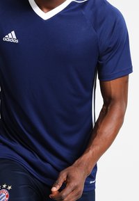 Marinblå idrottströja med V-ringning, vita detaljer och Adidas-logotyp. Tillverkad av andningsbart material, har korta ärmar och en åtsittande design.