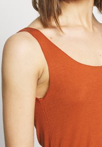 Top na ramiączkach w kolorze rdzy, o prążkowanej fakturze, szerokich ramiączkach, zaokrąglonym dekolcie i wykończeniu z niedokończonym szwem w pobliżu otworu na rękę.