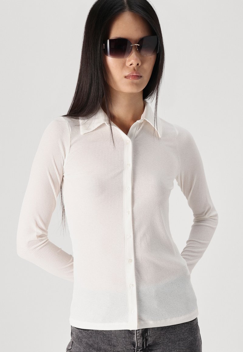 Langärmliges, tailliertes Hemd aus leichtbeigem geripptem Stoff, mit einem kragenlosen Ausschnitt und einer Knopfleiste. Kombiniert mit dunklen Sonnenbrillen.