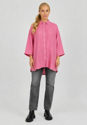Femme debout, portant une chemise ample rose à boutons, un jean gris droit et des bottines noires sur un fond clair uni.