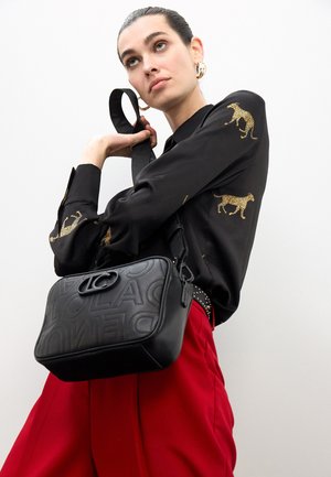 Bolso bandolera de cuero negro con detalle de logotipo en relieve, forma rectangular y correa ajustable; combinado con una camisa negra y pantalones rojos.