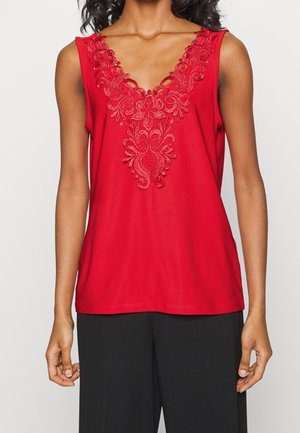 Kobieta w czerwonym, bezrękawniku z kwiatowym haftem koronkowym przy dekolcie w kształcie litery V, w połączeniu z czarnymi spodniami o szerokich nogawkach.