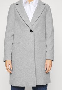 Person, der einen mittellangen grauen Wollmantel mit einem einzelnen schwarzen Knopf über einem weißen Hemd und blauen Jeans trägt.