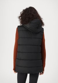 Doudoune noire sans manches avec sections matelassées horizontales et capuche, portée avec un pull côtelé orange, sur fond blanc.