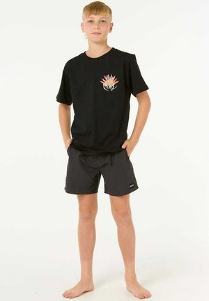 T-shirt nera in cotone con una grafica colorata e testo sul petto, abbinata a pantaloni da bagno neri. Il modello è in piedi a piedi nudi.