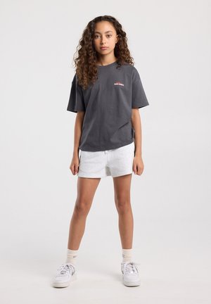 Jong persoon met krullend haar staat en draagt een donkergrijs Santa Monica T-shirt, lichtgrijze korte broek, witte sokken en witte sneakers tegen een witte achtergrond.