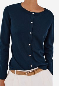 Tummansininen neulecardigan pitkillä hihoilla, pyöreällä kaula-aukolla ja hopeisella napitus edessä. Pehmeä, neulottu rakenne; käytetään beigen vyön päällä.