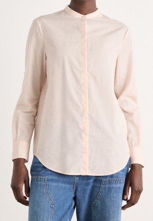 Personne portant une chemise beige clair boutonnée avec un col montant et un jean bleu, montrée des épaules aux hanches sur un fond uni.