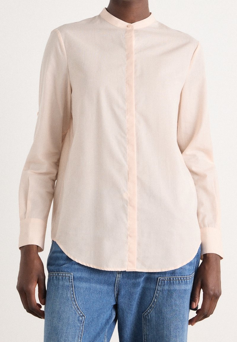 Personne portant une chemise beige clair boutonnée avec un col montant et un jean bleu, montrée des épaules aux hanches sur un fond uni.