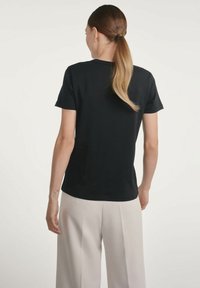 Czarna bawełniana koszulka z krótkim rękawem i okrągłym dekoltem. Widok z tyłu pokazuje gładką fakturę i dopasowany krój. Noszona z jasnymi spodniami.