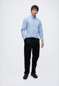 Junger Mann steht mit den Händen in den Taschen, trägt ein hellblaues Hemd mit Knöpfen, schwarze Hose und schwarze Schuhe vor weißem Hintergrund.