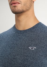 Mörkblå stickad tröja med rund hals och stickade muddar, med en liten broderad logotyp på bröstet. Mjuk textur, avslappnad design.