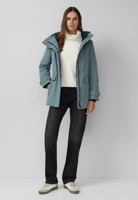 Žena stoji nasuprot sivog zida, odjevena u plavu jaknu s kapuljačom, bijeli pulover s visokim ovratnikom, crne hlače i svjetlosmeđe tenisice.