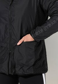 Veste noire présentant un tissu lisse d'un côté et un motif tissé texturé de l'autre. Zip sur le devant et poches latérales incluses.