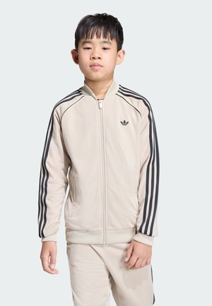 Béžová športová bunda s čiernymi pruhmi na rukávoch, so zipsom vpredu, rebrovanými manžetami a logom Adidas na hrudi. Vidieť aj zodpovedajúce nohavice.