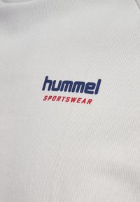 Close-up af hvidt stof med mørkeblå "hummel" og rød "SPORTSWEAR" trykte tekst.
