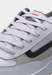 Λευκό αθλητικό παπούτσι με λευκά κορδόνια, μαύρη πλαϊνή ρίγα και γκρι και κόκκινες λεπτομέρειες στη γλώσσα, κοντινή εικόνα στην περιοχή των κορδονιών.