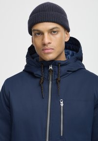 Navy blue hooded jacket with a zip front, adjustable drawstrings, and a chest pocket with a metallic zipper. Ribbed navy beanie.