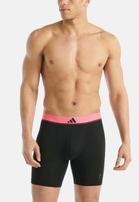 Spierkrachtige man in zwarte sportcompressiekorte broek met een roze tailleband en het Adidas-logo, staande tegen een witte achtergrond.