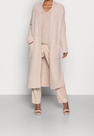 Frau trägt langen beigefarbenen, strukturierten Cardigan, passendes V-Ausschnitt-Oberteil, weit geschnittene Hosen und offene Sandalen, steht vor einer schlichten Wand.